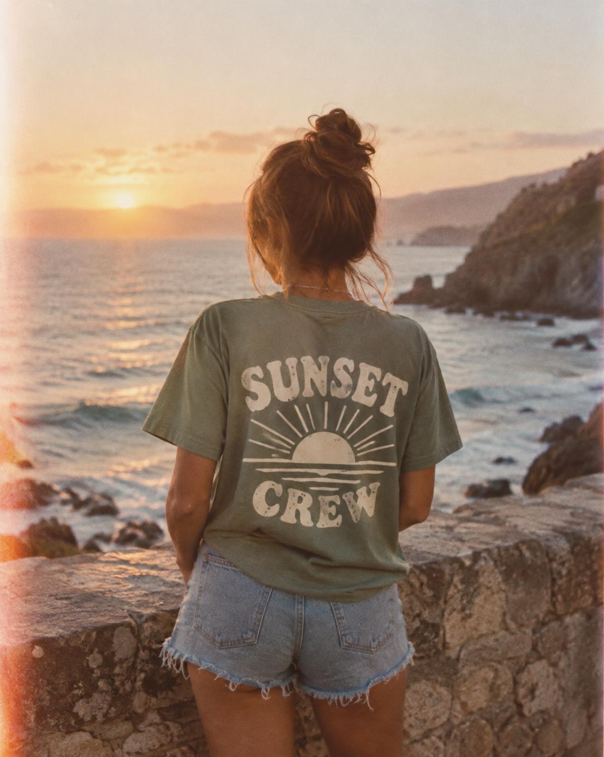 Woman at a seaside wall in a sage tee with a cream Sunset Crew print on the back